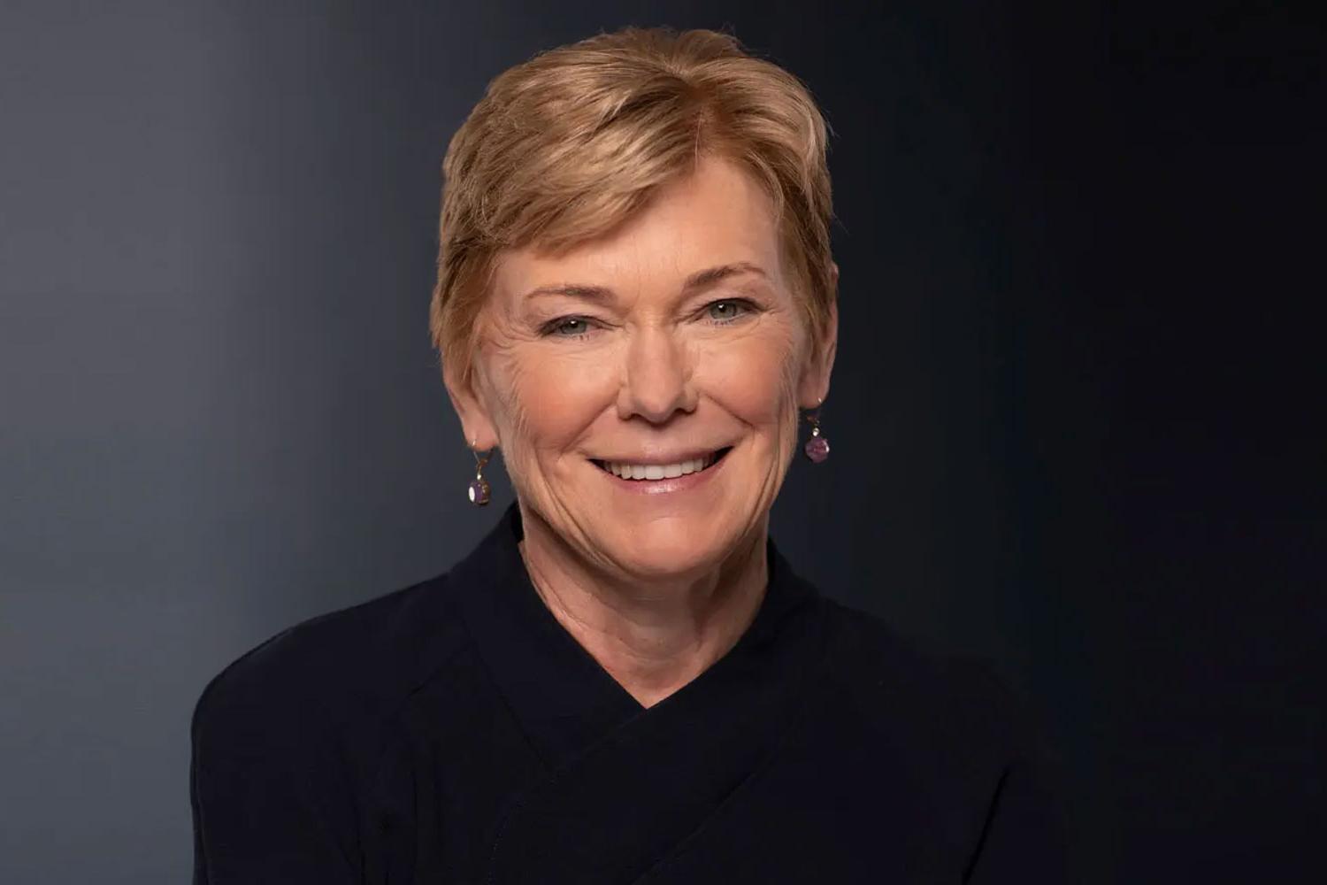 President Sarah C. Mangelsdorf, wearing a blue blazer and black shirt, stands in front of a bookshelf at the University of Rochester.